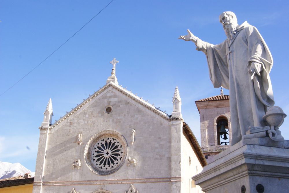 Statua di San Benedetto a Norcia, con la Basilica sullo sfondo, il rosone gotico e il campanile sotto un cielo limpido.