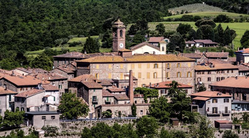 Borgo di Piegaro, immerso tra colline verdi e alberi, con edifici in pietra. In primo piano la ciminiera dell’antica vetreria