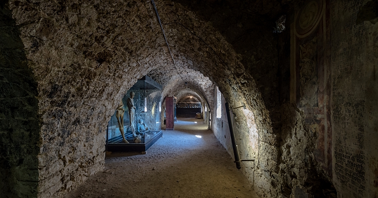 Un couloir souterrain avec des murs en pierre, des vitrines contenant des momies et de vieilles fresques délavées.
