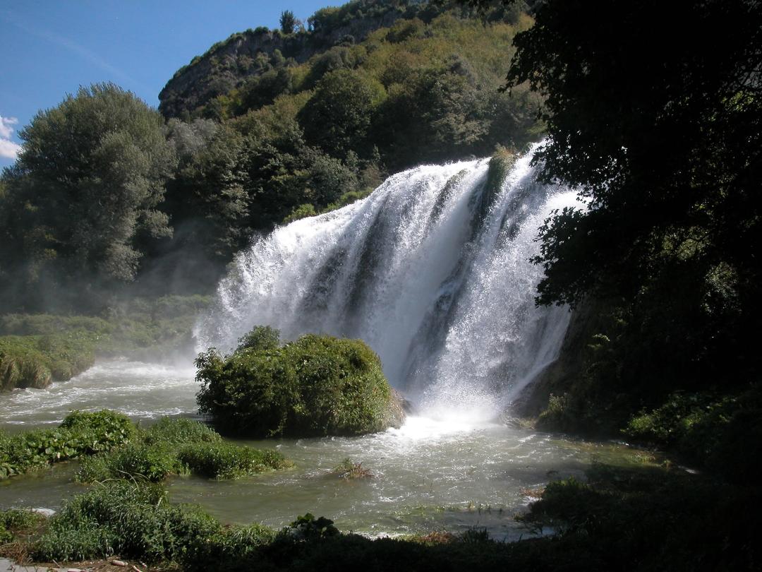The Marmore Waterfall seen from the side: water flows crashing against rocks, surrounded by lush vegetation and trees.