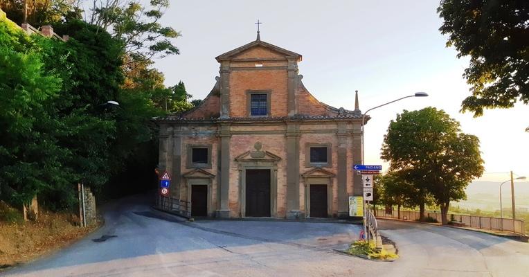 Una piccola Chiesa della Madonna della Sbarrain mattoni, circondata da alberi, si trova a un bivio con cartelli stradali.