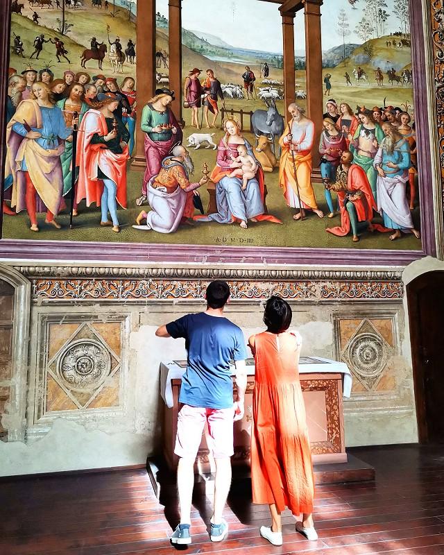Two people closely observe a Renaissance fresco depicting the Adoration of the Magi in a decorated church.