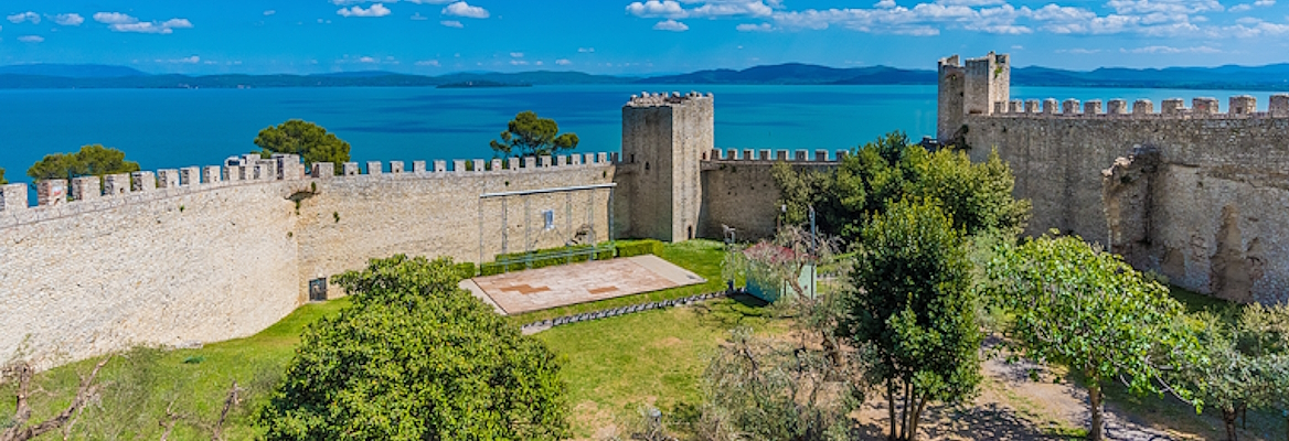 Panoramic view of the Rocca del Leone, with its imposing walls surrounding medieval towers and an open-air amphitheater inside. In the background, Lake Trasimeno and its islands create a picturesque scene.