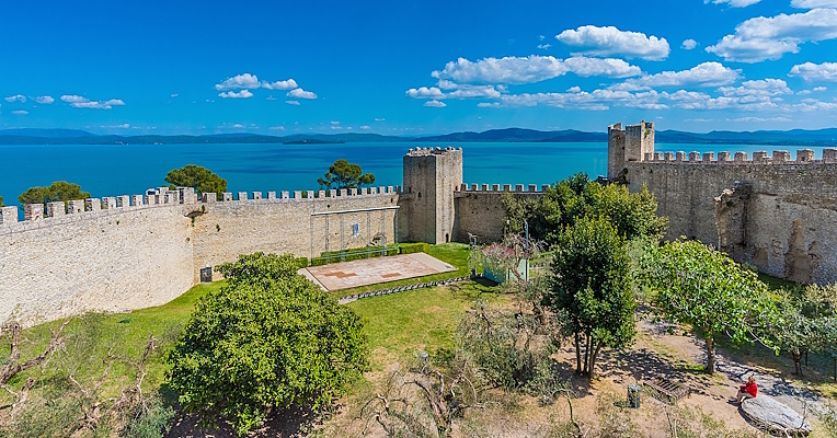  Panoramic view of the Rocca del Leone, with its imposing walls surrounding medieval towers and an open-air amphitheater inside. In the background, Lake Trasimeno and its islands create a picturesque scene. 
