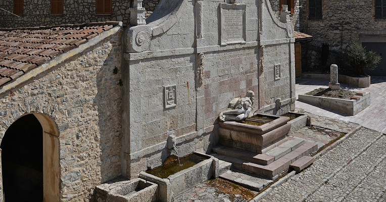 Fontaine en pierre avec décoration sculpturale et bassin dans un village de montagne, entouré de bâtiments anciens et d’un paysage vallonné.