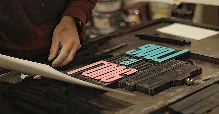 Artisan travaillant sur une impression typographique traditionnelle. Ses mains soulèvent une feuille de lettres roses et bleues pour former l’inscription « LOVE is LONG ».