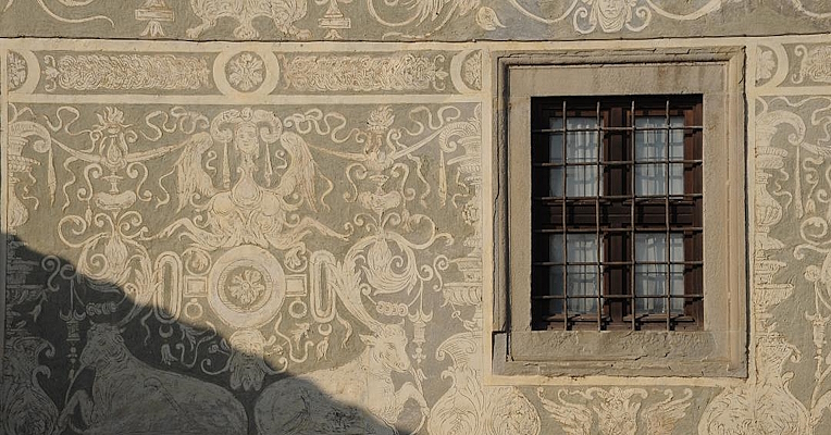 Mur décoré de motifs Renaissance blancs sur fond gris et fenêtre à barreaux au premier plan.