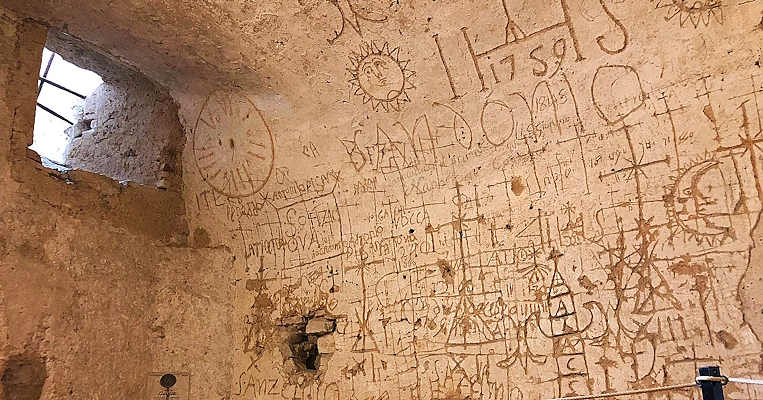 Walls engraved with symbols, crosses, and centuries-old inscriptions in a prison cell lit by a small barred window.