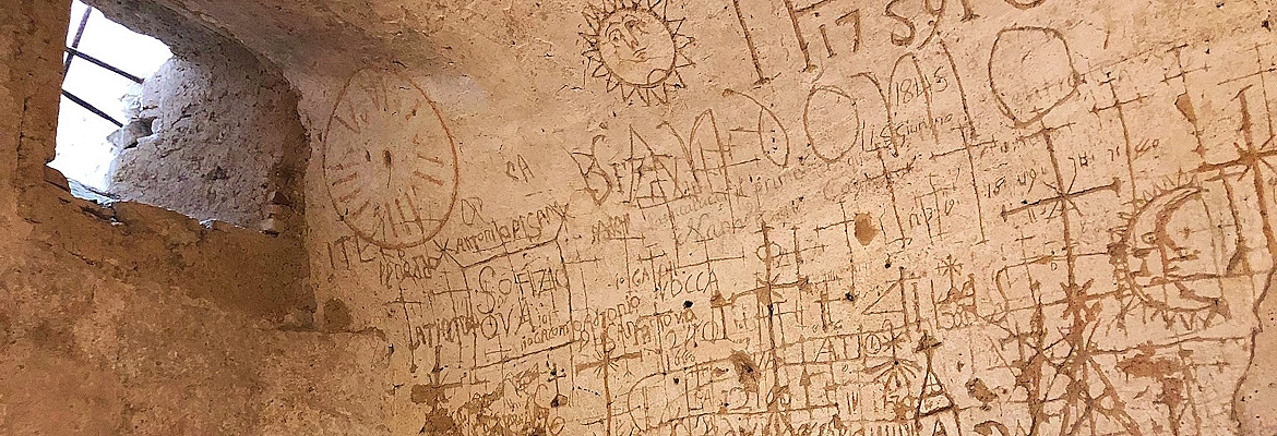 Walls engraved with symbols, crosses, and centuries-old inscriptions in a prison cell lit by a small barred window.