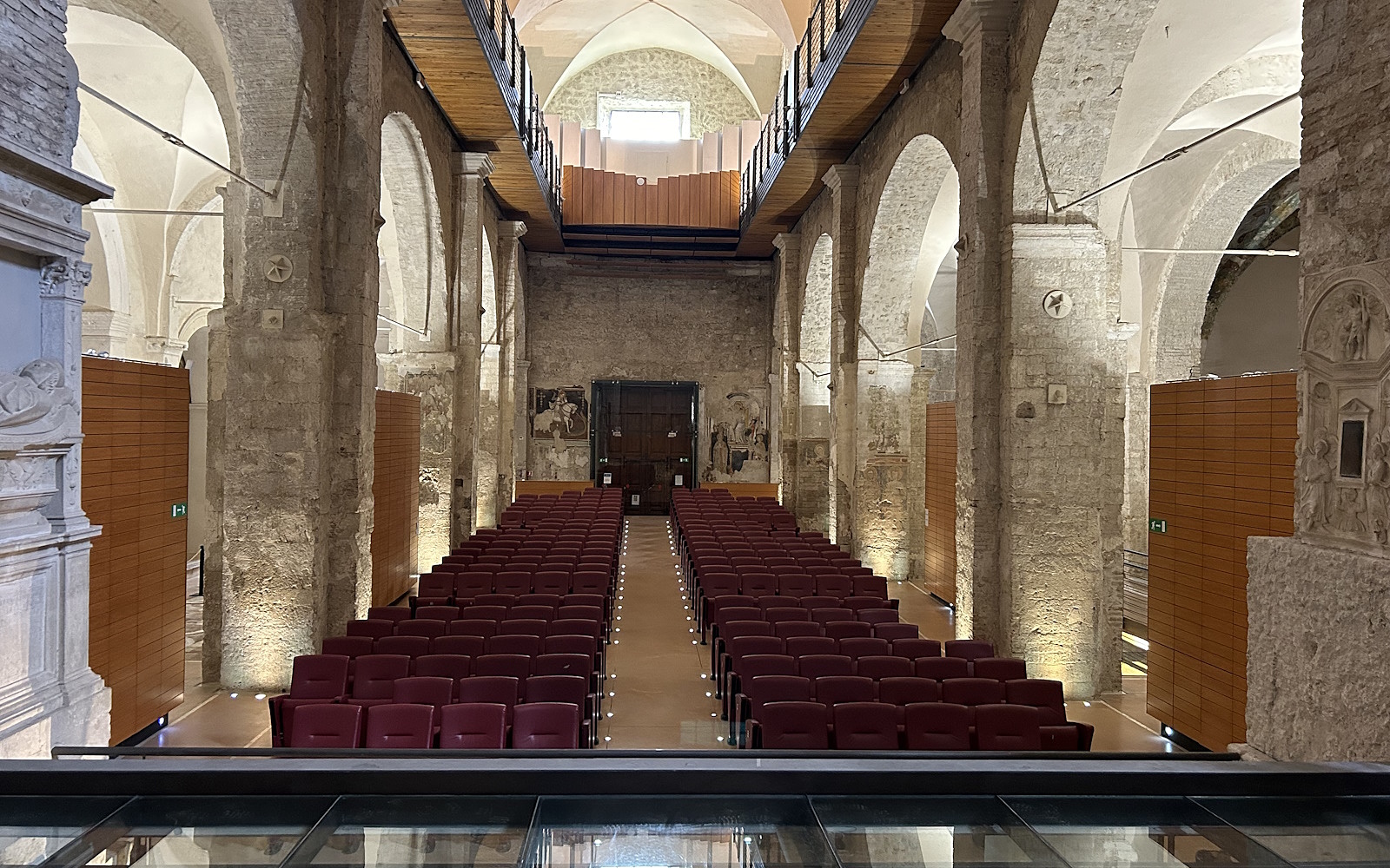Innenansicht des Bortolotti Auditoriums in Narni mit symmetrisch angeordneten Reihen roter Sitze, Steinwänden, einer Decke mit Kreuzgewölben und einem hölzernen Laufsteg auf der oberen Etage, der sich entlang beider Seiten des Saals erstreckt. Im Hintergrund schmücken eine große Holztür und teilweise sichtbare Fresken die Wand.