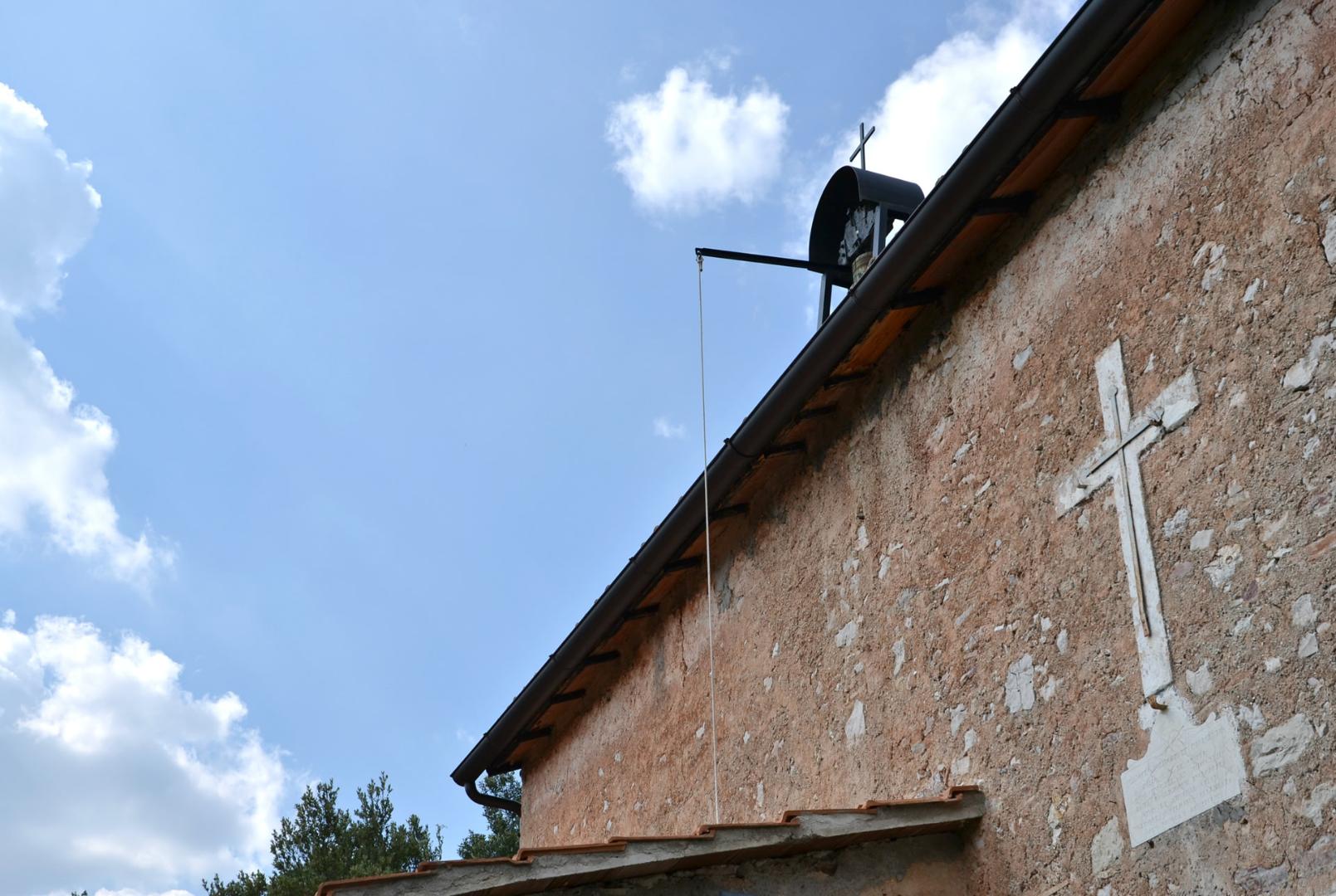 Stone façade of a small church with white cross and bell gable under a blue sky dotted with clouds.