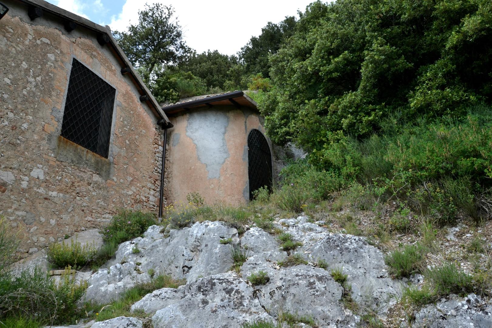 Stone exterior of the Hermitage of Madonna del Riparo, with walls and barred windows, set among greenery and rocks.