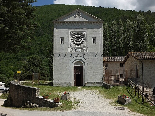 Romanesque white stone church surrounded by greenery, with central rose window and arched portal, nestled among wooded hills.