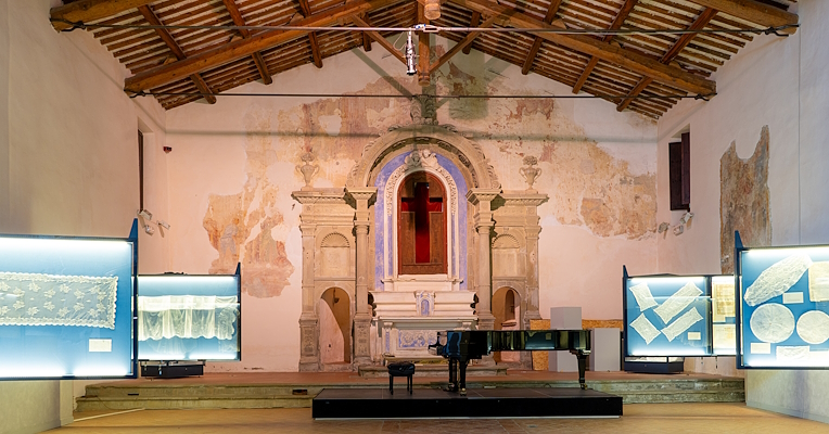 Intérieur de l'église, désormais siège du musée, avec des vitrines exposant des dentelles et un autel en arrière-plan. Au centre, un piano.