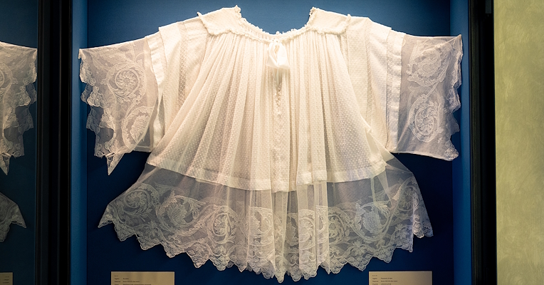 Robe de baptême blanche brodée en tulle, avec des manches larges et des détails décoratifs, exposée dans une vitrine du musée.