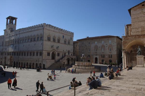 Piazza IV Novembre con fontana maggiore e palazzo dei priori, passanti si godono la giornata soleggiata seduti sulle scale del duomo o passeggiando.