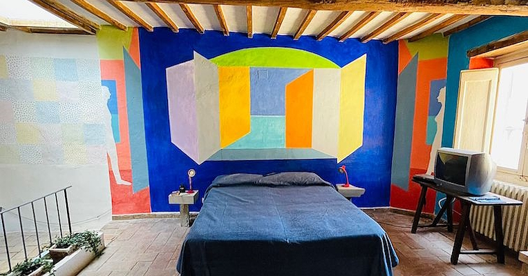 Schlafzimmer mit blauer Wand, die mit farbigen geometrischen Formen hinter dem Bett verziert ist, und freiliegenden Balken an der Decke.