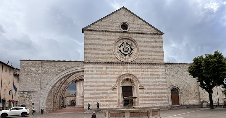 Façade de la Basilique de Santa Chiara à Assise, avec rosace et arc latéral, vue depuis la place avec fontaine au premier plan.