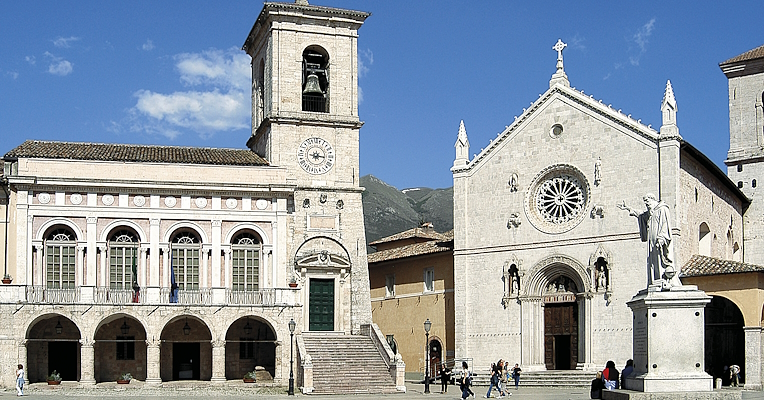 Place centrale avec le Palais Municipal et la Basilique de San Benedetto à Norcia, avec un clocher et une statue au premier plan.