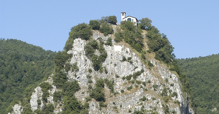 Éperon rocheux avec végétation et une petite église au sommet, entouré d'un paysage de collines vertes sous un ciel clair.