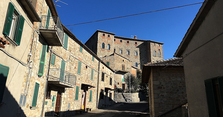 Rue du village de Carnaiola avec maisons en pierre et volets verts, sous un ciel clair; un château en haut de la rue.