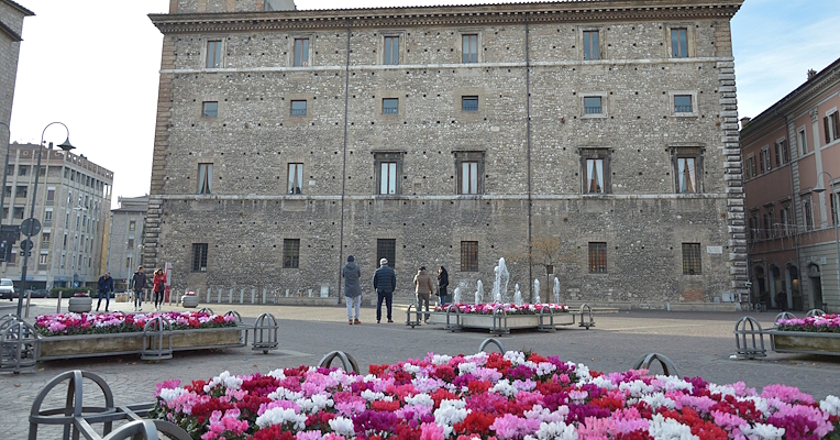 Piazza della Repubblica à Terni, avec des parterres de cyclamens roses, rouges et blancs, tandis que des passants observent le Palazzo Spada