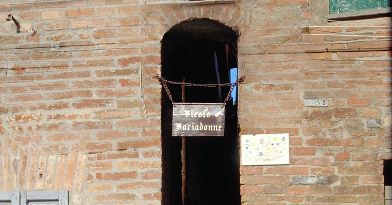 Ruelle étroite entre des murs de briques, avec un panneau marron suspendu par des chaînes indiquant en blanc : « Vicolo Baciadonne ».