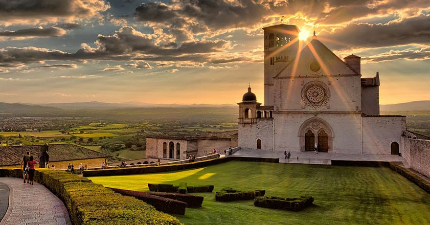 La basilique Saint François d’Assise au coucher du soleil, le soleil filtrant à travers les nuages et illuminant la prairie et la vallée.