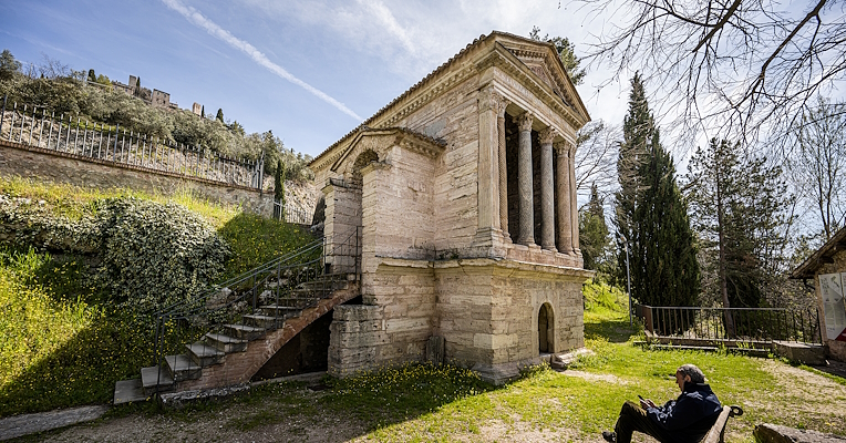 Aspetto esterno del Tempietto sul Clitunno, con quattro colonne e la scalinata laterale. In primo piano, un uomo seduto su una panchina