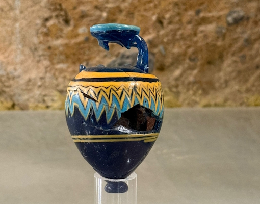 Ancien balsamaire en verre brisé au col et au ventre, avec des motifs décoratifs bleus et jaunes sur fond bleu, exposé sur un support transparent.