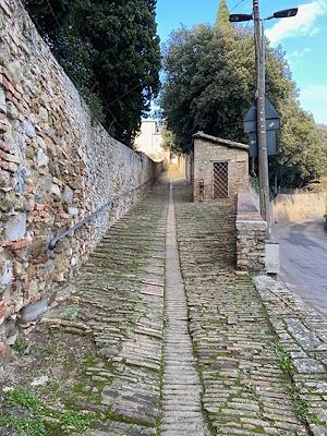 Escalier en pierre et en briques avec un petit mur latéral, bordé d’un mur en pierre et d’arbres, menant à un couvent au fond.