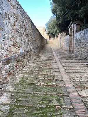 Escalier escarpé en pierre et en briques, flanqué de murs et d’arbres, menant à un couvent au fond sous un ciel clair.