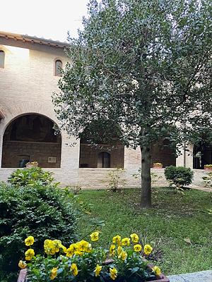Cloître avec un portique en briques, un jardin bien entretenu avec des fleurs jaunes et un arbre au centre, dans une atmosphère sereine et historique.