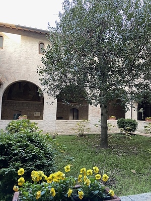 Ein Kreuzgang mit einem Backsteinportikus, ein gepflegter Garten mit gelben Blumen und einem Baum in der Mitte, in einer ruhigen und historischen Atmosphäre.