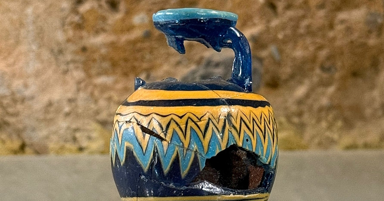  Ancient glass paste unguentarium with a fractured neck and body, featuring decorative blue and yellow motifs on a blue background, displayed on a transparent stand 