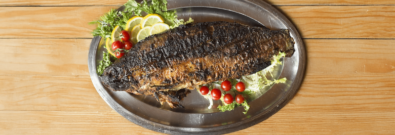 Immagine: Carp in porchetta on a steel tray, garnished with lemon, salad, and cherry tomatoes on a wooden table. 