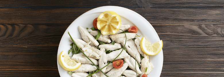 Immagine: Perch fillets served with rocket, cherry tomatoes, chives, and lemon slices on a white plate, placed on a wooden table. 