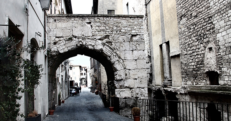 Roman stone arch in a narrow street in Spoleto, with historic buildings on either side preserving remnants of Roman columns