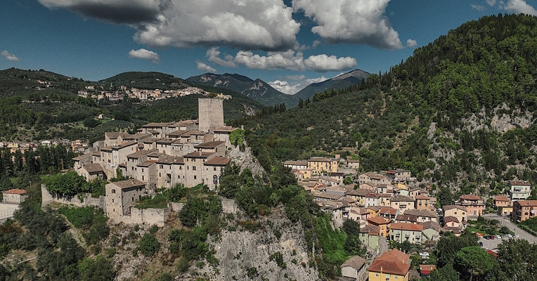 Veduta aerea di un borgo medievale su uno sperone roccioso, tra colline e montagne circostanti.