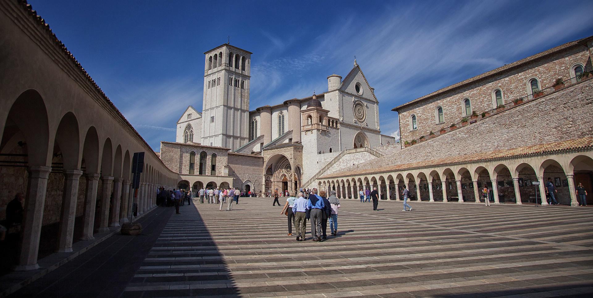 Piazza Inferiore di San Francesco in Assisi, gekennzeichnet durch steinerne Seitenportale und die Präsenz der Oberen Basilika im Hintergrund. Die zentrale Treppe führt zum Eingang der Oberen Basilika und unterstreicht die gotische Architektur des Platzes.