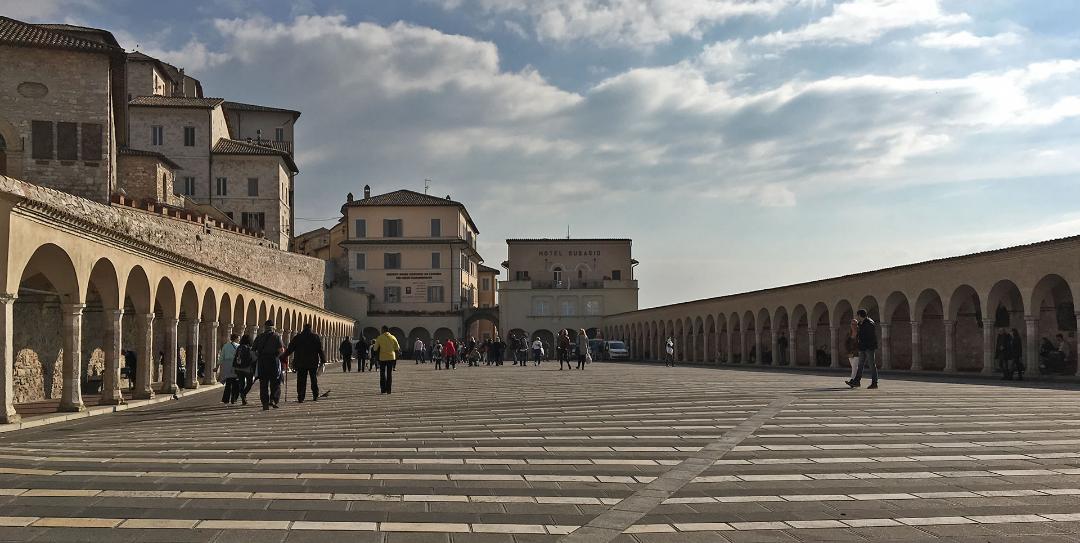 Blick auf den Platz vor der Basilika des Heiligen Franziskus in Assisi, der durch breite steinerne Säulengänge auf beiden Seiten gekennzeichnet ist.