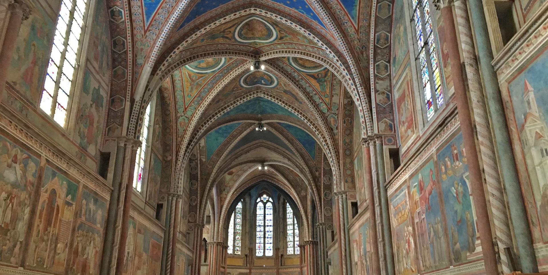 Innenansicht der Oberen Basilika des Heiligen Franziskus in Assisi mit ihren hohen, verzierten Kreuzgewölben. Die Wände, die mit Fresken von Künstlern wie Giotto und Cimabue bedeckt sind, zeigen Szenen aus dem Leben des Heiligen Franziskus und biblische Geschichten. Große Fenster lassen natürliches Licht in den Innenraum einfallen.