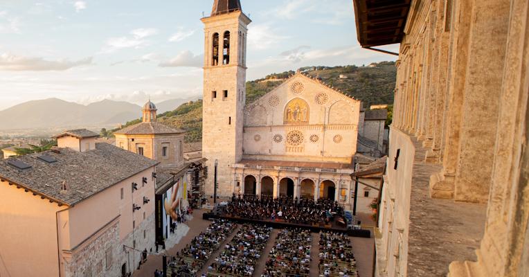 High-angle view of Piazza Duomo in Spoleto during an evening of the Festival dei Due Mondi, with the audience seated in front of the stage set up in front of the Romanesque cathedral, illuminated by the warm light of sunset, with the Umbrian hills and the town below in the background.