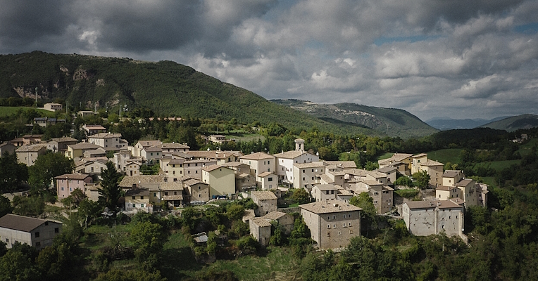 Aerial view of Poggiodomo, with the historic centre surrounded by green hills under a cloudy sky.