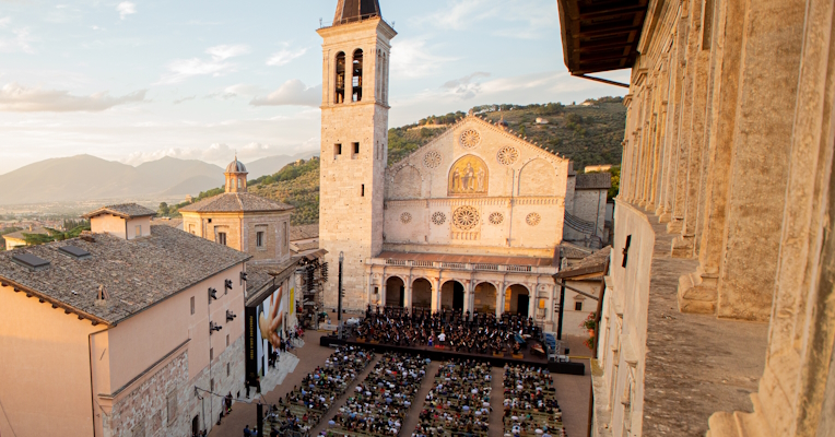  High-angle view of Piazza Duomo in Spoleto during an evening of the Festival dei Due Mondi, with the audience seated in front of the stage set up in front of the Romanesque cathedral, illuminated by the warm light of sunset, with the Umbrian hills and the town below in the background. 