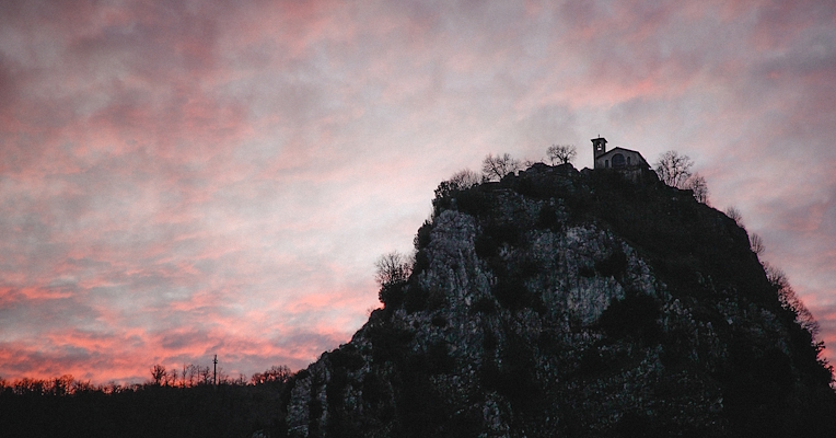 Silhouette of the Sanctuary of Roccaporena at sunset, perched atop the rock, with pink sky and streaked clouds.