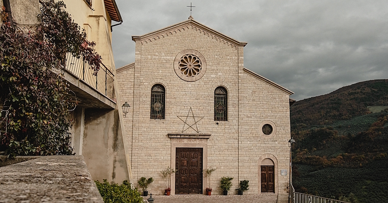 Stone façade of Santa Maria Assunta in Montefranco, with rose window, wooden door, and Christmas decorations.