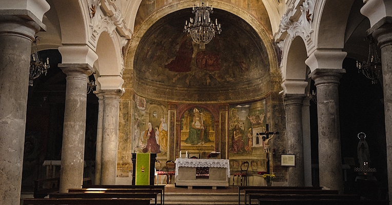 Interior of the Church of San Nicola in Scheggino, with stone columns, frescoed altar and hanging chandeliers.