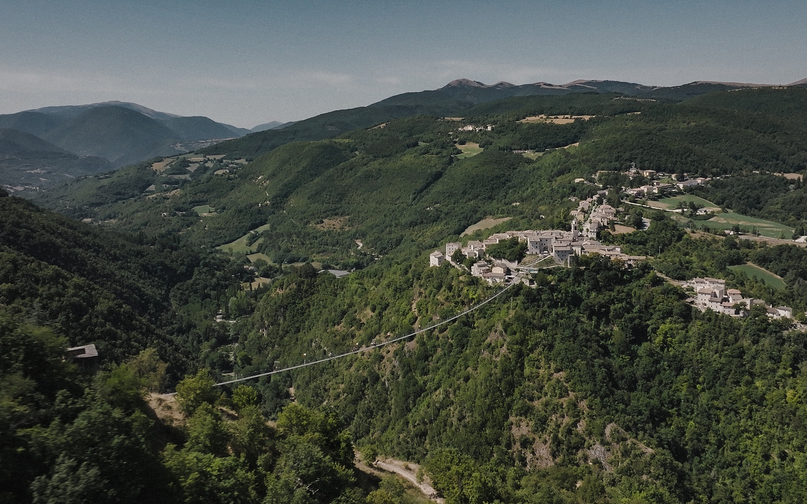  View of the Sellano Tibetan bridge suspended between two wooded hills, with the village perched and the valley in the background. 