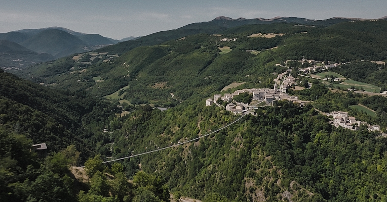 View of the Sellano Tibetan bridge suspended between two wooded hills, with the village perched and the valley in the background.